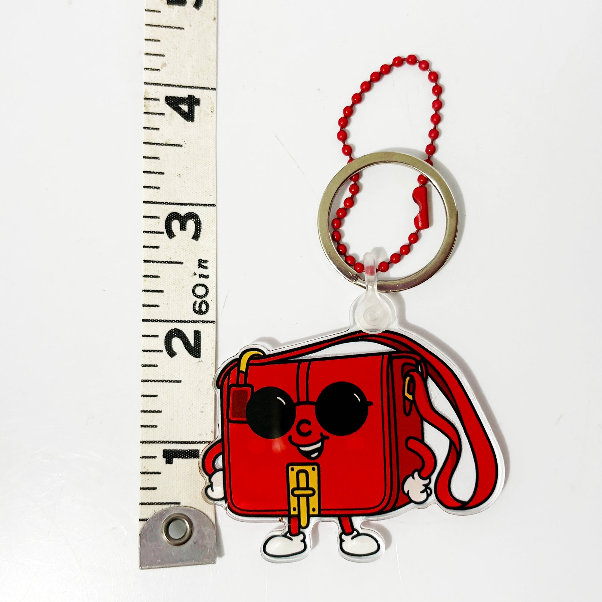 Red cartoon-style purse keychain bag charm with sunglasses on a white background, next to a ruler for scale.