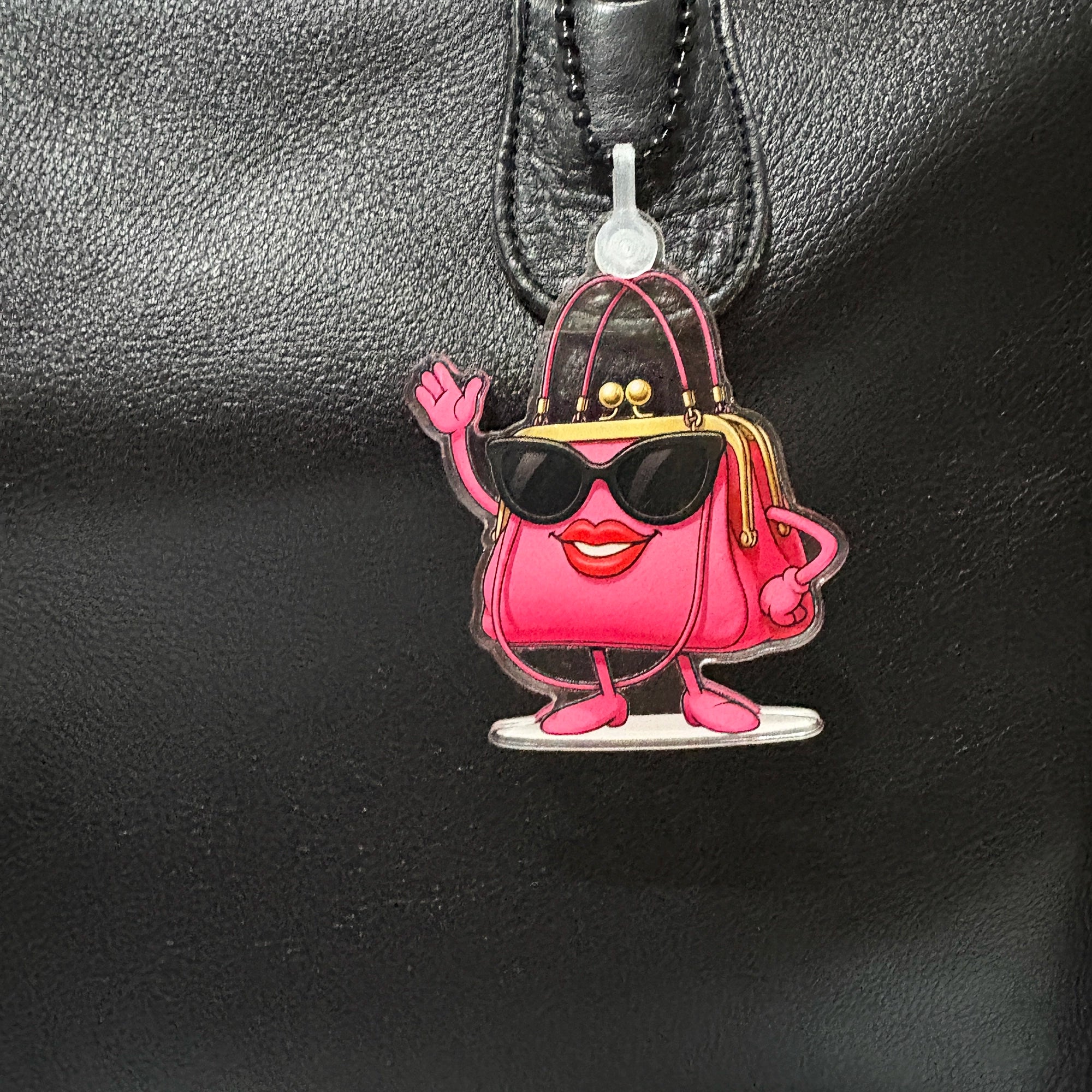 Sassy Pink cartoon-style handbag charm with sunglasses and a smiley face on a black leather surface.